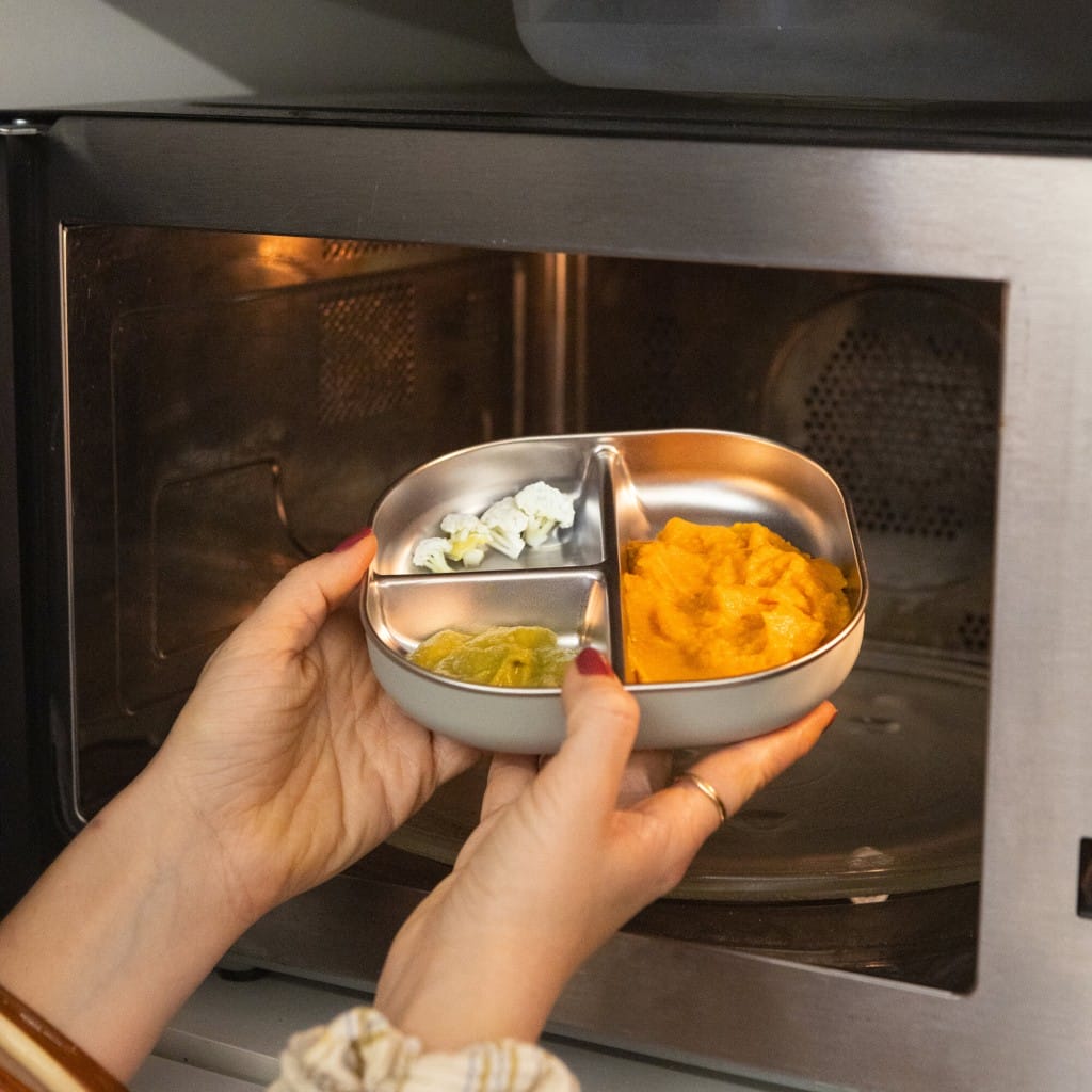 Adult placing food in a Cotton Candy Beaba divided plate into a microwave